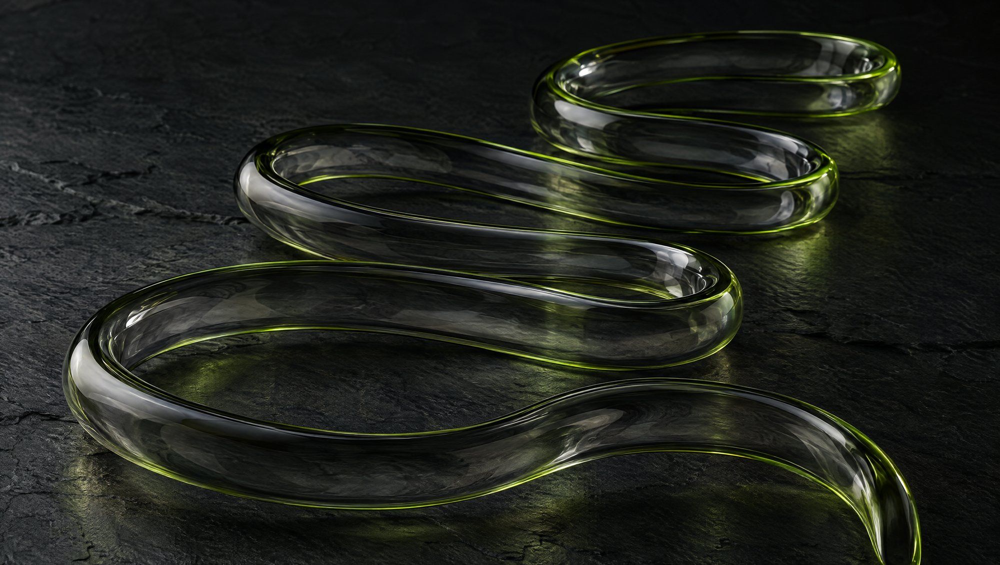 A long clear glass tube coiled in loose curves on a dark slate surface, its edges lit in olive green.