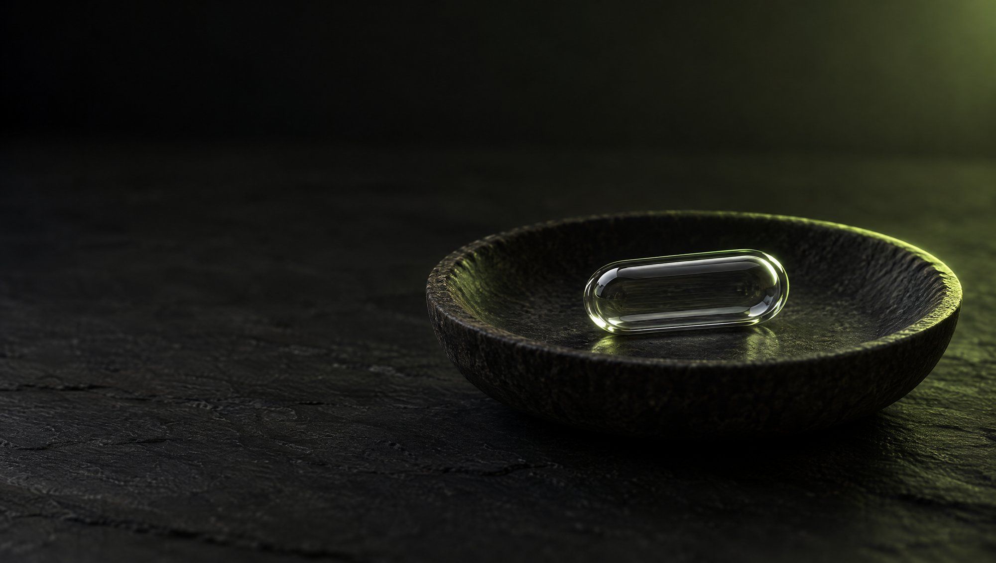 A single clear glass capsule resting in a shallow dark stone dish on a slate surface, lit from behind with olive-green light.