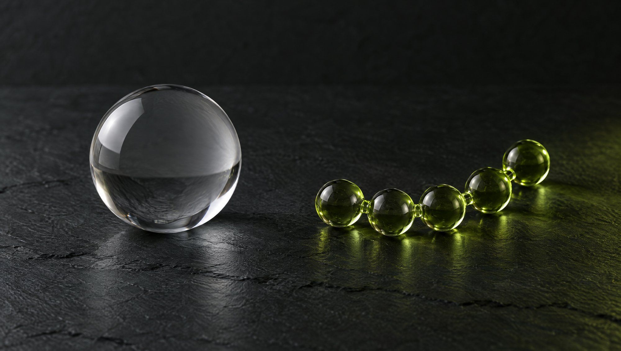 A clear glass sphere in neutral light beside a short chain of small olive-green glass beads on a dark slate surface.
