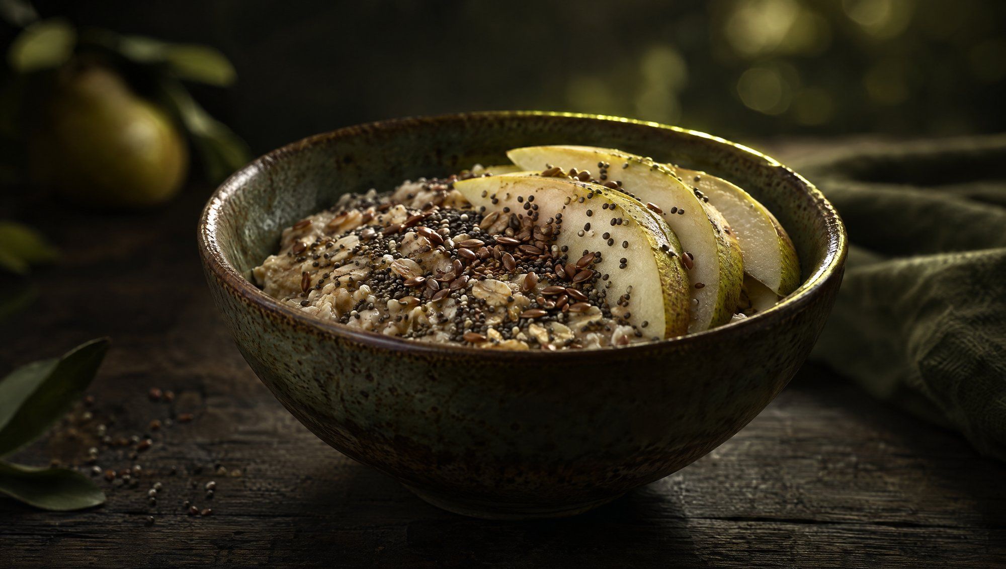 A rustic ceramic bowl of rolled oats topped with chia seeds, flax seeds and thin slices of pear on a dark wooden table, lit from behind with olive-green light against soft olive bokeh.
